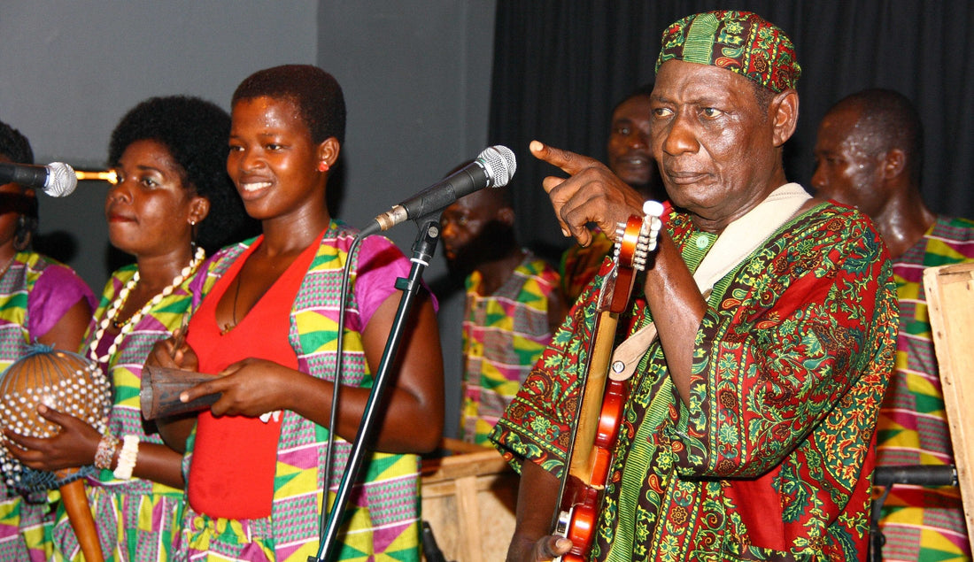 A highlife band on stage, the bandleader holding a guitar and pointing off-mic, singers beside him with a shaker and a gong-gong