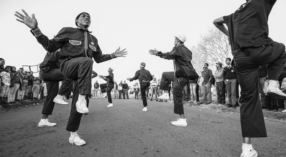 Pantsula dancers in a circle on a tarred township street, knees high, spectators ringed around them, shot in black and white.